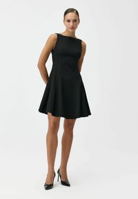 Kobieta stoi, ubrana w czarną, dopasowaną sukienkę bez rękawów o kroju rozkloszowanym oraz czarne wysokie obcasy na jednolitym białym tle.