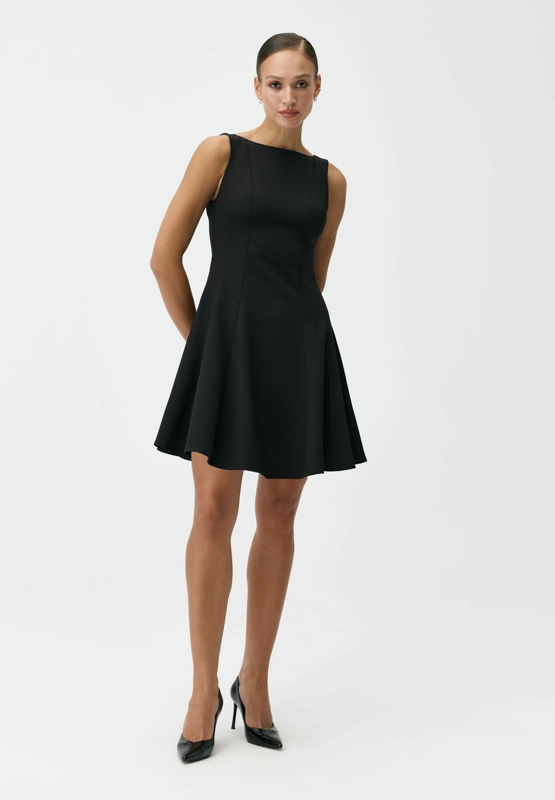 Kobieta stoi, ubrana w czarną, dopasowaną sukienkę bez rękawów o kroju rozkloszowanym oraz czarne wysokie obcasy na jednolitym białym tle.