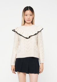 Cream knitted sweater with a black-trimmed ruffled collar, featuring a textured pattern and ribbed cuffs, paired with a dark pleated skirt.