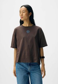 Bruine katoenen t-shirt met een relaxte pasvorm, ronde hals en blauwe bloemenprint in het midden. Gecombineerd met blauwe denim jeans.