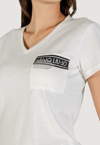 Camiseta blanca con cuello en V que presenta un bolsillo negro con un diseño y logo metálico. Tela suave y ligera con un ajuste casual.