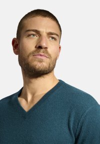 Svetr s výstřihem do V v tmavě modrozelené pletenině, s jemnou texturou a přiléhavým designem, který zvýrazňuje výstřih a horní část trupu.