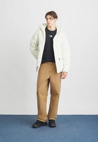 Blouson en duvet blanc avec capuche, t-shirt noir avec logo, pantalon taille haute marron, et chaussures noires, se tenant sur un tapis bleu devant un fond gris.