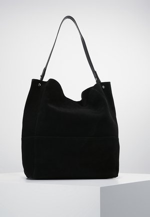 Sac cabas en daim noir avec une bandoulière unique, une structure souple et décontractée, et des détails minimums en rivets argentés sur un socle blanc.