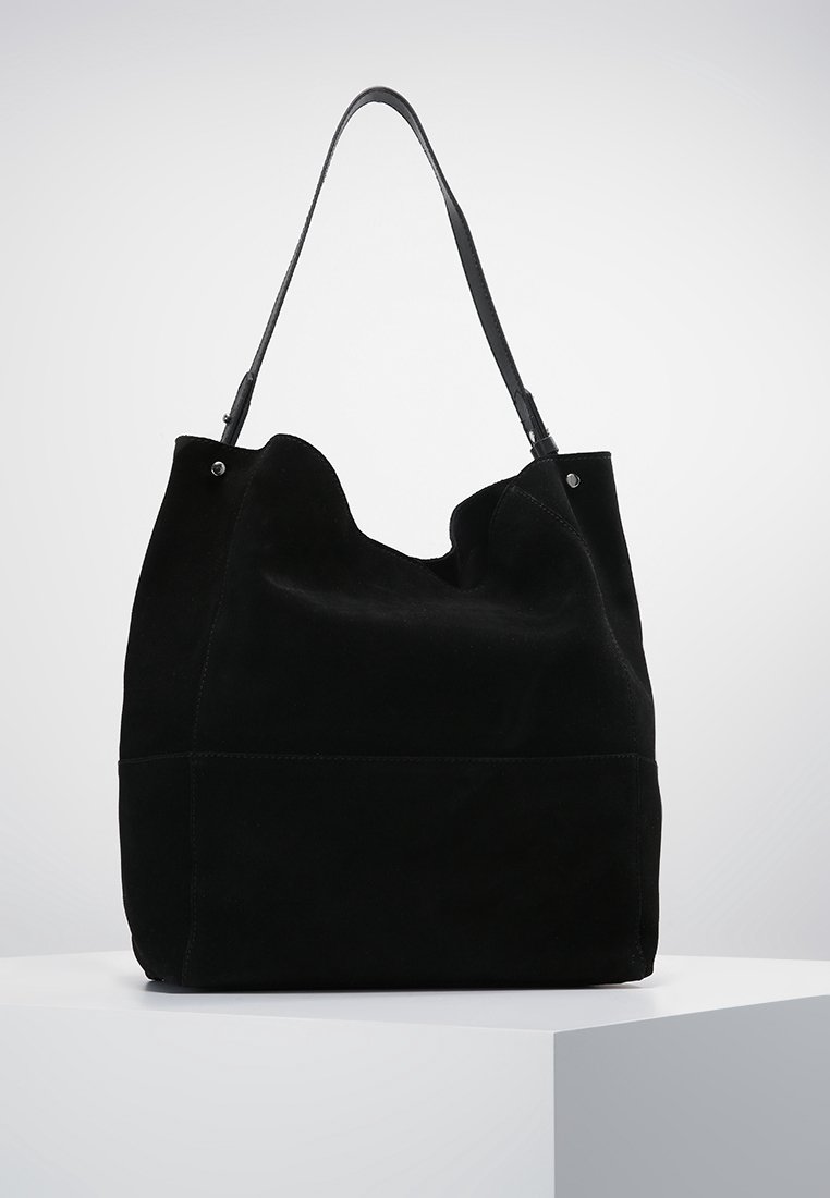Sac cabas en daim noir avec une bandoulière unique, une structure souple et décontractée, et des détails minimums en rivets argentés sur un socle blanc.