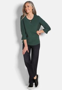 Lächelnde Frau mit langen grauen Haaren posiert in einem dunkelgrünen 3/4-Ärmel-Oberteil, schwarzen Hosen und schwarzen Turnschuhen mit weißen Sohlen.