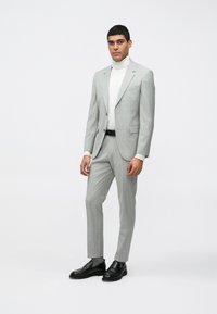 Fato cinza claro com um blazer estruturado e calças slim-fit, combinado com um suéter branco de gola alta e sapatos pretos. Tecido suave, design minimalista.