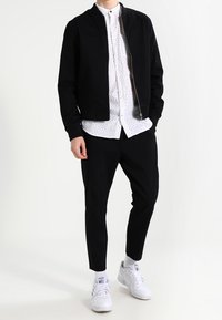 Svart bomberjacka över en vit skjorta med ljus mönstring, i kombination med svarta tapered byxor och vita sneakers.