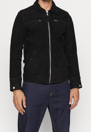 Homme portant une veste zippée en suède noire avec poches latérales et un jean bleu foncé, debout devant un fond clair uni.