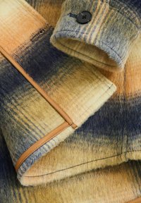 Tejido de lana a cuadros en naranja, amarillo y azul; presenta un zipper marrón y un botón negro en el puño; textura suave con un aspecto cálido.