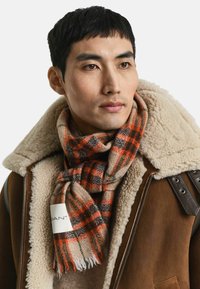 Écharpe en laine à carreaux orange, marron et beige, avec des bords fringés. Portée avec une veste en shearling marron, étiquette visible avec le logo.