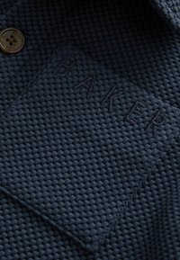 Marineblauwe textielstof met diamantpatroon, met de geborduurde tekst "BAKER" en een metalen knoopdetail.