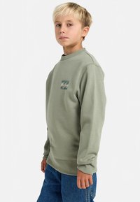 Ragazzo biondo con un felpa verde chiaro con un logo a onda sul petto e jeans blu, in piedi contro uno sfondo semplice.