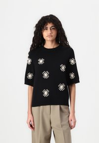 Maglione nero a maniche corte con ricami floreali crema, collo e orlo a coste, abbinato a pantaloni chiari e morbidi.