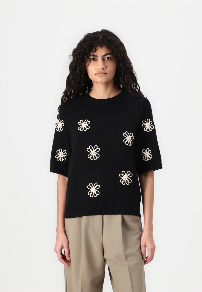 Maglione nero a maniche corte con ricami floreali crema, collo e orlo a coste, abbinato a pantaloni chiari e morbidi.