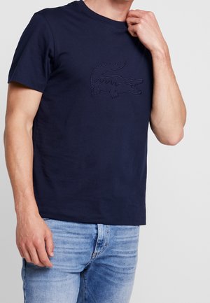 T-shirt en coton bleu marine avec un logo crocodile texturé en noir. Col rond et manches courtes. Porté avec un jean bleu clair.