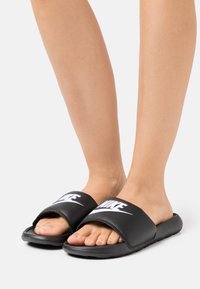 Fekete csúszó szandál széles pánttal, fehér "NIKE" logóval, lapos talppal és puha textúrájú anyaggal a kényelem érdekében.