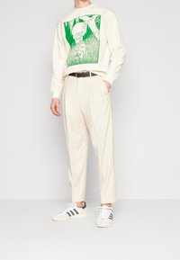 Kremowy sweatshirt z zieloną grafiką, zestawiony z jasnobeżowymi spodniami w kant. Obuwie to białe i szare sneakersy w czarne paski.