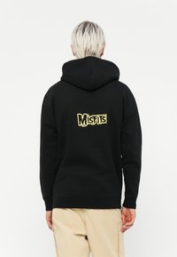 Black hoodie with a yellow embroidered "Misfits" logo on the back. Features a drawstring hood and ribbed cuffs and hem.