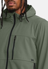 Chaqueta verde con capucha y cremallera, con detalles en negro y un logo. Tejido suave con una textura lisa, diseñada para proporcionar calidez y comodidad.