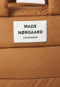 Pruunist pehmest kangast valmistatud kott, millel on valge silt, kus on musta tekstiga "MADS NØRGAARD COPENHAGEN". Sile tekstuur, ristkülikukujuline kuju.