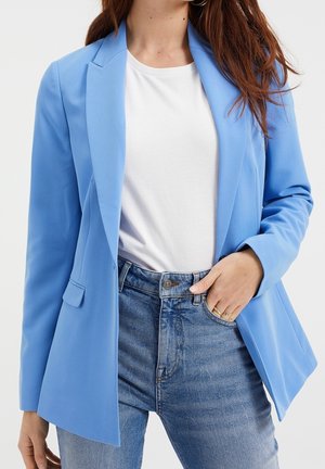 Vrouw draagt een lichtblauw blazer over een wit t-shirt met hoog getailleerde blauwe jeans, houdt één hand in de zak en ringen zijn zichtbaar.