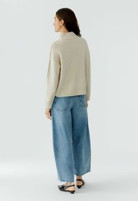 Beige gebreide trui met een hoge kraag, gecombineerd met lichtblauwe denim jeans met wijde pijpen. De jeans hebben achterzakken en een ontspannen pasvorm.