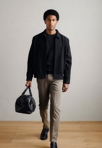 Musta takki, musta neule, beiget housut, mustat kengät, kädessään teksturoitu musta nahkalaukku, kävelee puulattialla neutraalia seinää vasten.