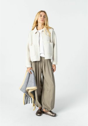 Frau mit langen blonden Haaren, die eine weiße Jacke und lockere beige Hose trägt und in der linken Hand eine grau-gelbe Fransen-Decke hält.