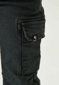 Pantalones cargo negros con una tela texturizada, que presentan bolsillos laterales y un pequeño logotipo en la parte delantera. Los detalles incluyen costuras cosidas y cierres seguros.