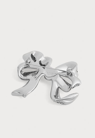 Broche en métal argenté en forme de nœud stylisé avec une fermeture à épingle à l'arrière sur un fond clair uni.