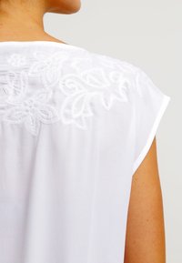 Weiße Bluse mit floralem Stickerei auf dem Rücken, aus weichem Stoff, kurzen Ärmeln und einer lockeren Passform, die zarte gestickte Details präsentiert.