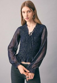 Mornarsko bluza z teksturiranim smockanom korpusom, volančastim izrezom in prosojnimi dolgimi rokavi. Zaveže se spredaj; subtilne metalne niti dodajajo podrobnosti.