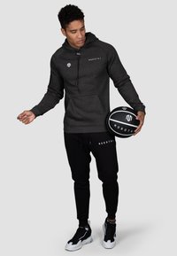 Dunkelgrauer Kapuzenpullover mit Logo, Fronttasche, schwarze Jogginghose mit Logo und schwarz-weißen Sneakers, hält einen schwarzen Basketball.