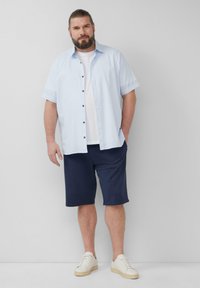 Lichtblauw kortemouwen button-up shirt over een wit T-shirt, gecombineerd met marineblauwe shorts en witte sneakers, tegen een effen achtergrond.