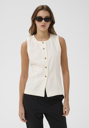 Femme portant un haut blanc sans manches boutonné et un pantalon noir, avec des cheveux bruns mi-longs et des lunettes de soleil noires œil de chat.