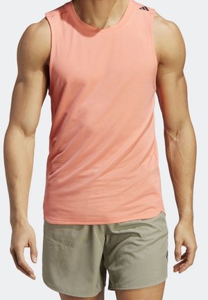 Muskelöser Mensch, der ein ärmelloses korallrosa Sporttop und hellgraue Shorts trägt und mit entspannten Armen an den Seiten steht.