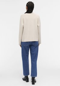 Beige gebreide trui met een ronde halslijn, lange mouwen en een losse fit, gecombineerd met blauwe hoge spijkerbroeken en zwarte blokhaklaarzen.