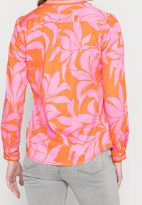 Blouse à manches longues avec un motif floral orange et rose, présentant une texture lisse, des poignets à boutons et un ourlet arrondi.
