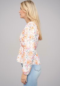 Blusa a stampa floreale bianca, con fiori arancioni e rosa, maniche lunghe con polsini elasticizzati e orlo a peplum. Abbinata a jeans azzurri chiari.