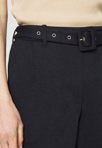 Svarta strukturerade byxor med ett matchande bälte med fyrkantigt spänne, metalöglor och sidfickor, sedd från midjan och ner.