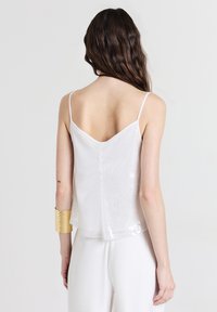 Woman with long brown hair wearing white sleeveless sequin top, white pants, and gold cuff bracelet, shown from the back on gray background.
