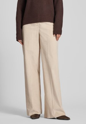 Pantaloni a gamba larga in corduroy beige con superficie texture, caratterizzati da un'alta vita e pieghe frontali, abbinati a un maglione in maglia marrone.