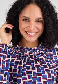 Blusa con scollatura arricciata, caratterizzata da un motivo astratto nei toni del blu, rosso e crema. Tessuto morbido, maniche lunghe e vestibilità rilassata.