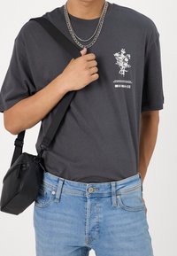 Grau Baumwoll-T-Shirt mit floralem Design und weißem Grafikdruck auf der linken Brust, kombiniert mit hellblauen Jeans und einer schwarzen Umhängetasche.