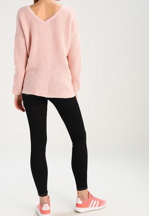 Maglione lavorato a maglia rosa chiaro con design a V sul retro, abbinato a leggings neri e scarpe da ginnastica rosa. Tessuto texture con una vestibilità rilassata.