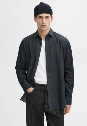 Camicia button-up in denim scuro con maniche lunghe, colletto a punta e taglio dritto, abbinata a jeans neri e un berretto di lana nero.