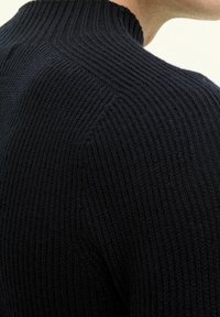 Svart stickad tröja med ribbad struktur och hög krage, som visar en åtsittande design som följer konturerna av halsen och axlarna.