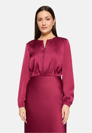 Frau mit langen braunen Haaren, die ein langärmliges, reißverschlossenes, tiefrosa Satinoberteil und einen passenden Rock vor einem einfarbigen Hintergrund trägt.