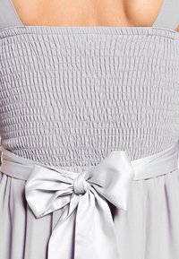 Světlé šedé šaty se smockovaným živůtkem a širokými ramínky. V oblasti pasu mají saténovou mašli, která zvýrazňuje texturovaný vzhled látky.